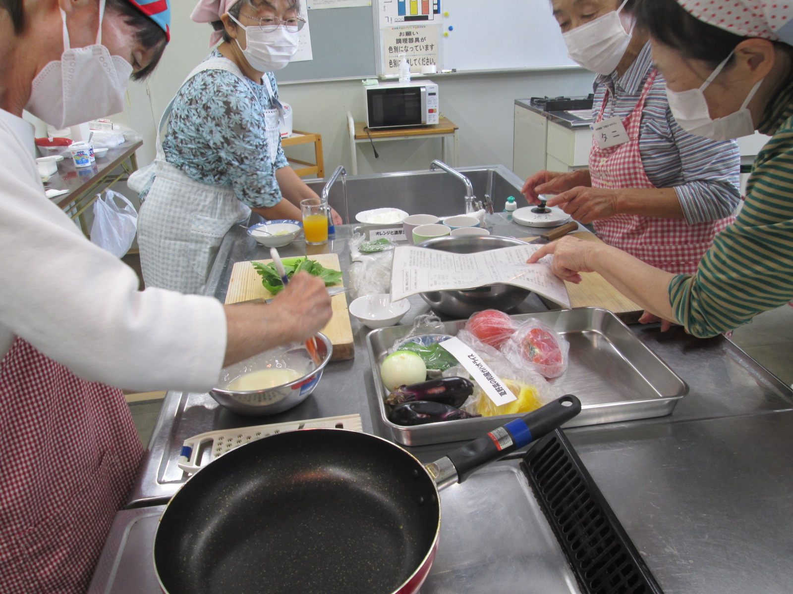 エプロンとマスクを着用した複数の参加者が、ステンレス製の調理台を囲み、野菜やフライパン、レシピを手に取りながら料理教室に参加している様子の写真