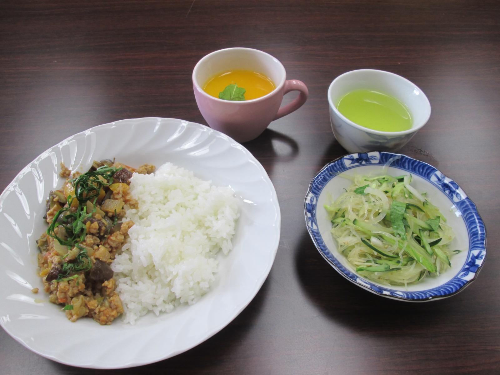 茶色のテーブルに並べられた、白いご飯と炒め物が盛られた白い皿、青い柄の小鉢に入った野菜サラダ、ピンクのマグカップに入ったスープ、白い小鉢に入った緑茶などが並べられている写真