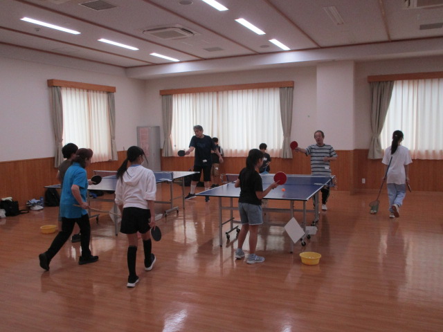 広い体育館のような室内で、2台の卓球台が設置されており、子どもから大人まで様々な年齢層の人がラケットを持って卓球を楽しんでいる様子を捉えた写真