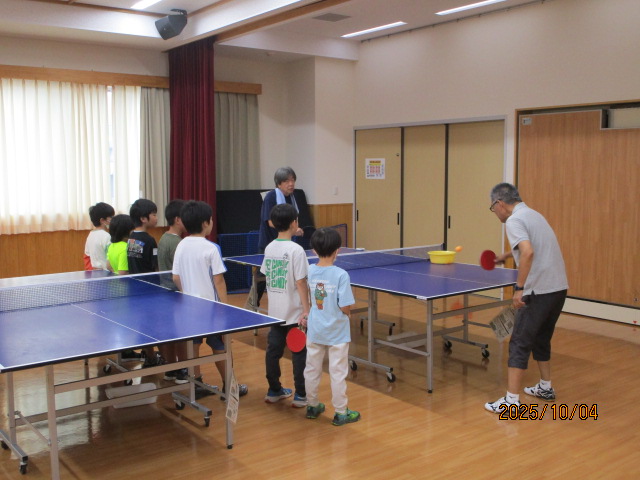 体育館のような室内の卓球台で、大人男性2人がラリーをしているところを子供たちが眺めている写真