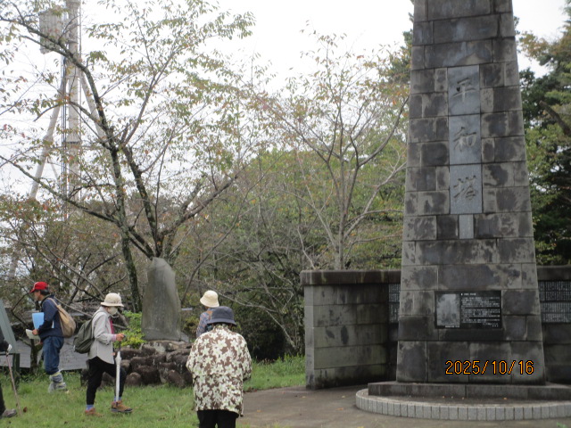 森の木々を背景に、大きな石積みの塔のような記念碑が立っており、数人の参加者がその周辺に集まって石碑を眺めている様子を捉えた写真