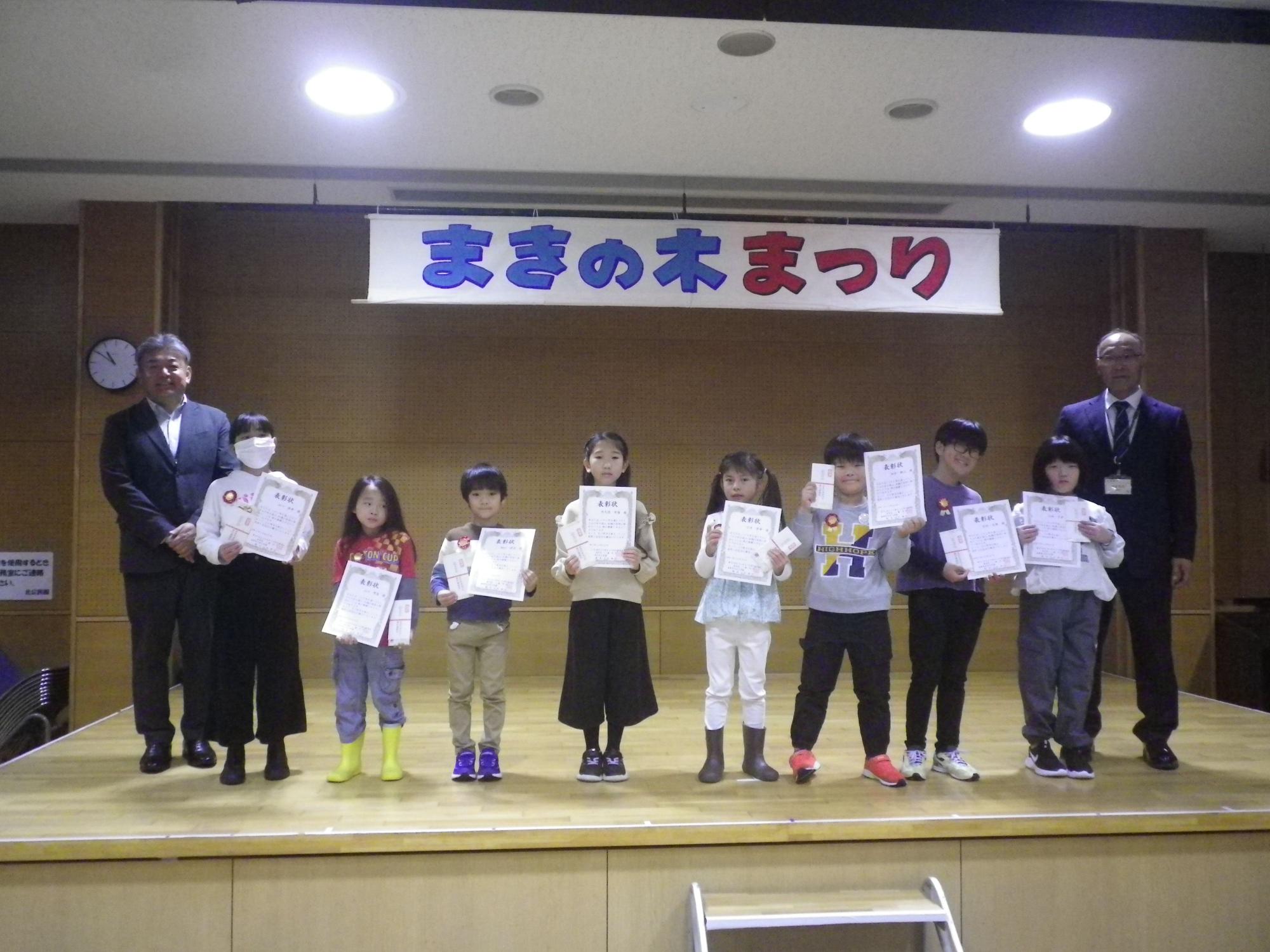まきの木まつりの横断幕の下、ステージ上で賞状を持った8人の子どもたちとスーツ姿の大人2人が並んで記念撮影している表彰式の写真