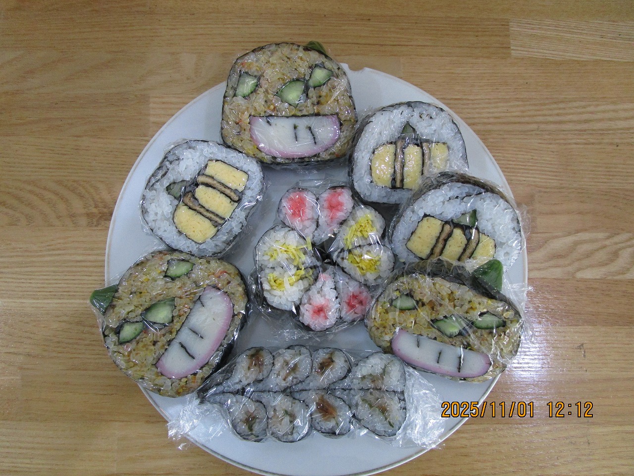 「かぼちゃ」や「お化けカボチャ」顔の形に海苔や卵、キュウリで装飾された巻きずしと藤の花をイメージした、賑やかな飾り巻き寿司を上から撮影した写真