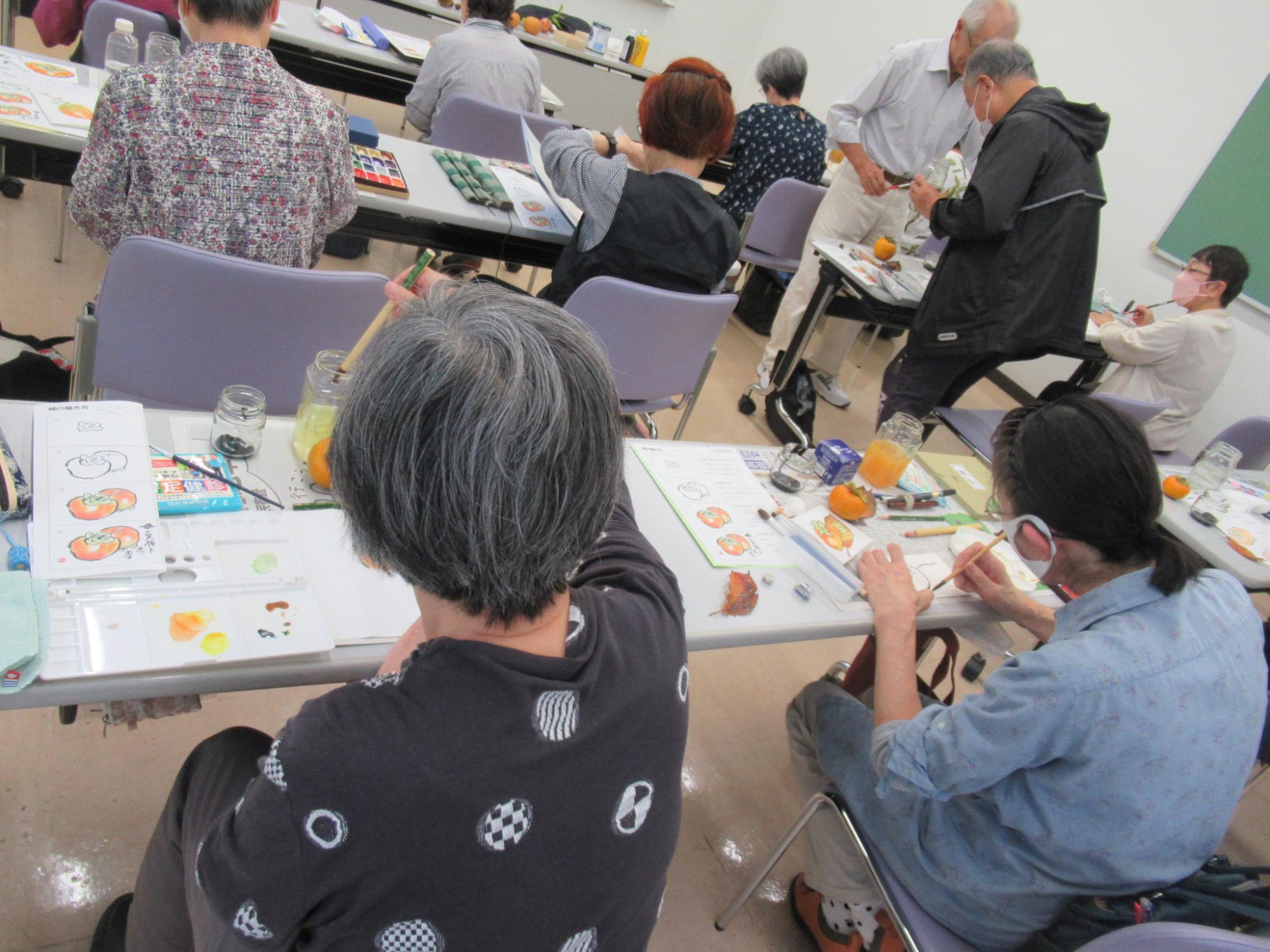 明るい室内で、数人の高齢者が長テーブルを囲み、手前で二人のお年寄りが筆と水彩絵の具を使って、卓上に置かれた本物の柿と見本の紙を見ながらスケッチや絵付けに集中して取り組んでいる様子を、後方から捉えたワークショップの様子の写真