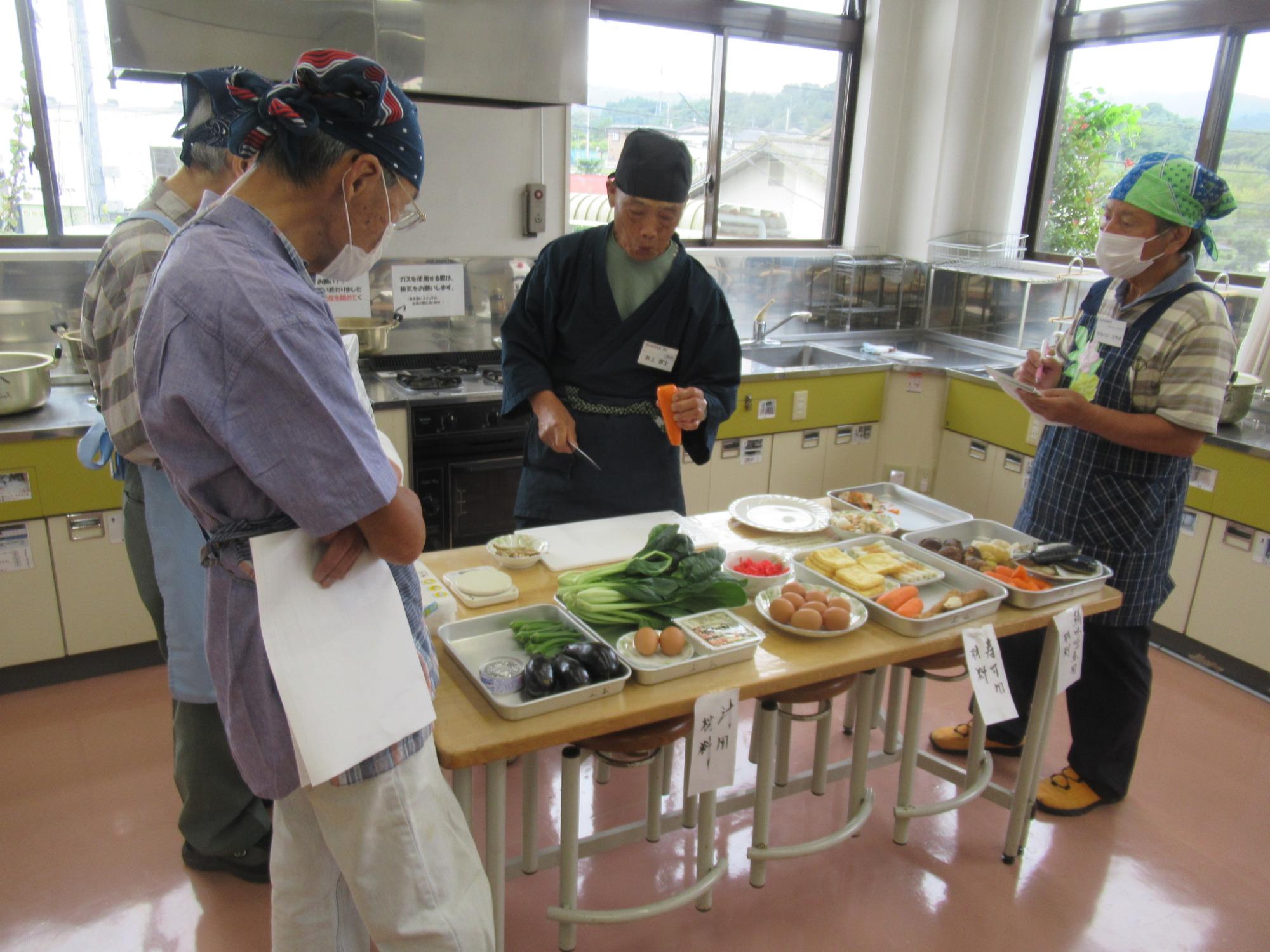 明るい調理実習室のような空間で、頭にバンダナや手ぬぐいを巻いた四人の高齢男性が、中央の作業台に並べられた野菜や卵などの食材を囲み、一人の男性が手に持った人参を切りながら説明をし、他の三人がメモを取ったり熱心に聞いたりしている様子の写真