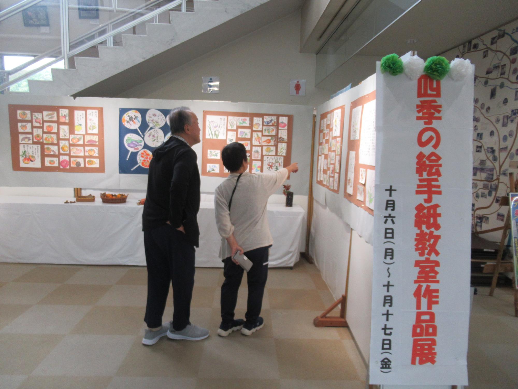 「四季の絵手紙教室作品展」と開催日程が書かれた縦長の白い案内板が立てられている広い屋内スペースで、二人の高齢者が壁に展示された水彩画や貼り絵のような数多くの作品を熱心に鑑賞している写真