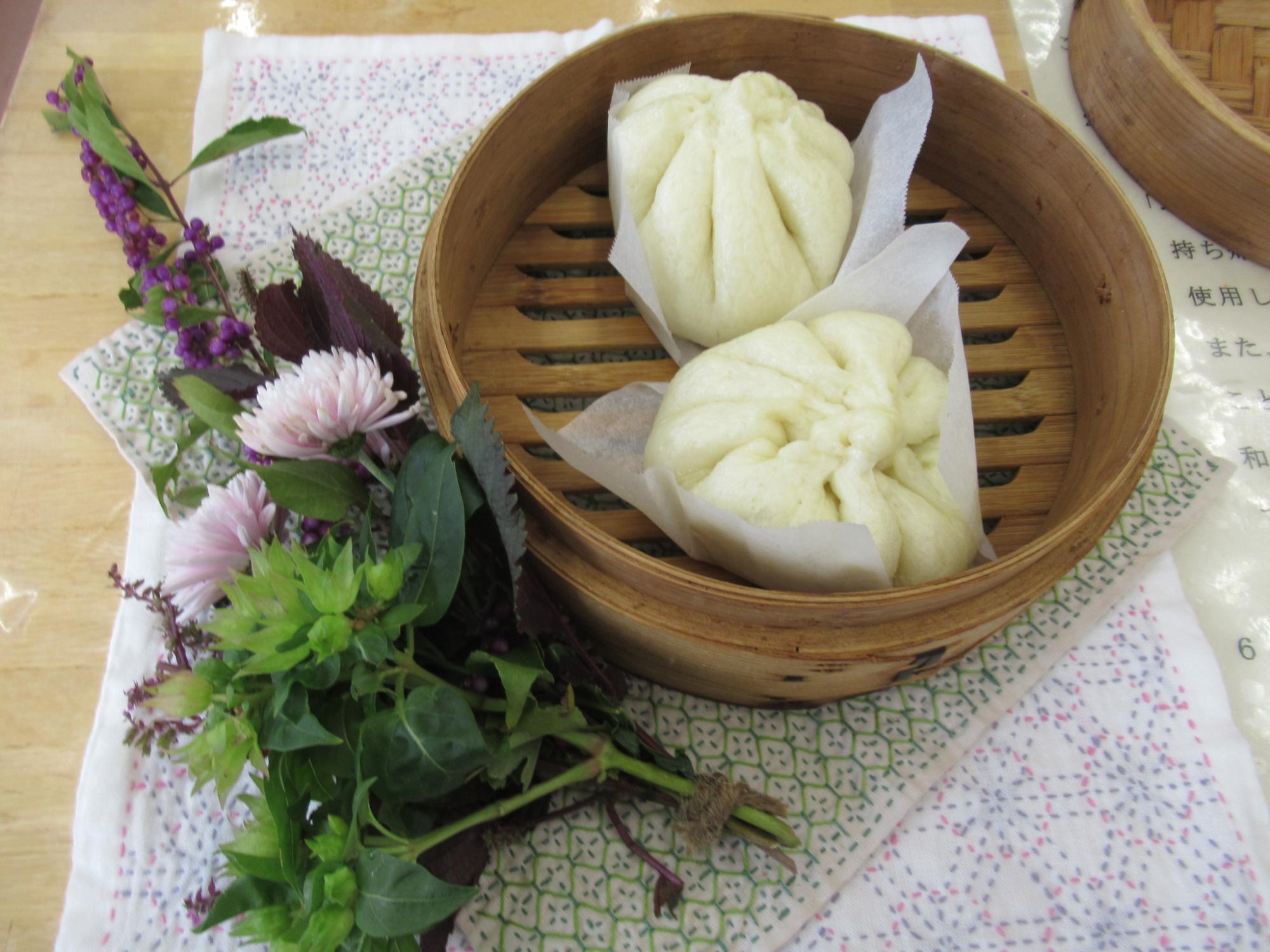 手前の左下には紫色の花と白い菊のような花、青々とした葉を束ねた生け花が添えられた、木製の蒸篭の中に敷かれた白いクッキングシートの上で蒸し上がったばかりの丸く白い肉まんが二つ並べられている写真