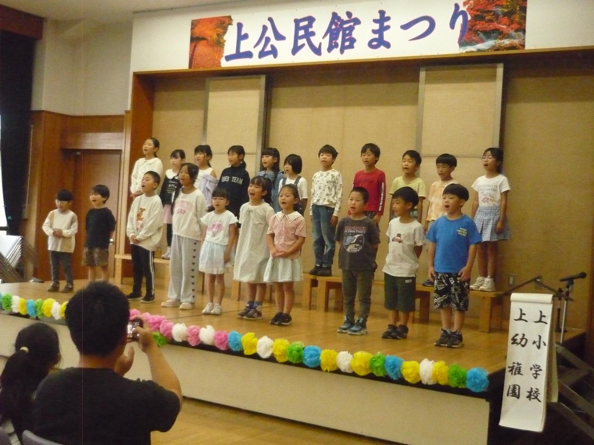 「上公民館まつり」と書かれた横断幕の下のカラフルなポンポン飾りが並んだステージに、大勢の子どもたちが2列で並んで歌を歌い、手前では観客がその様子を撮影している写真