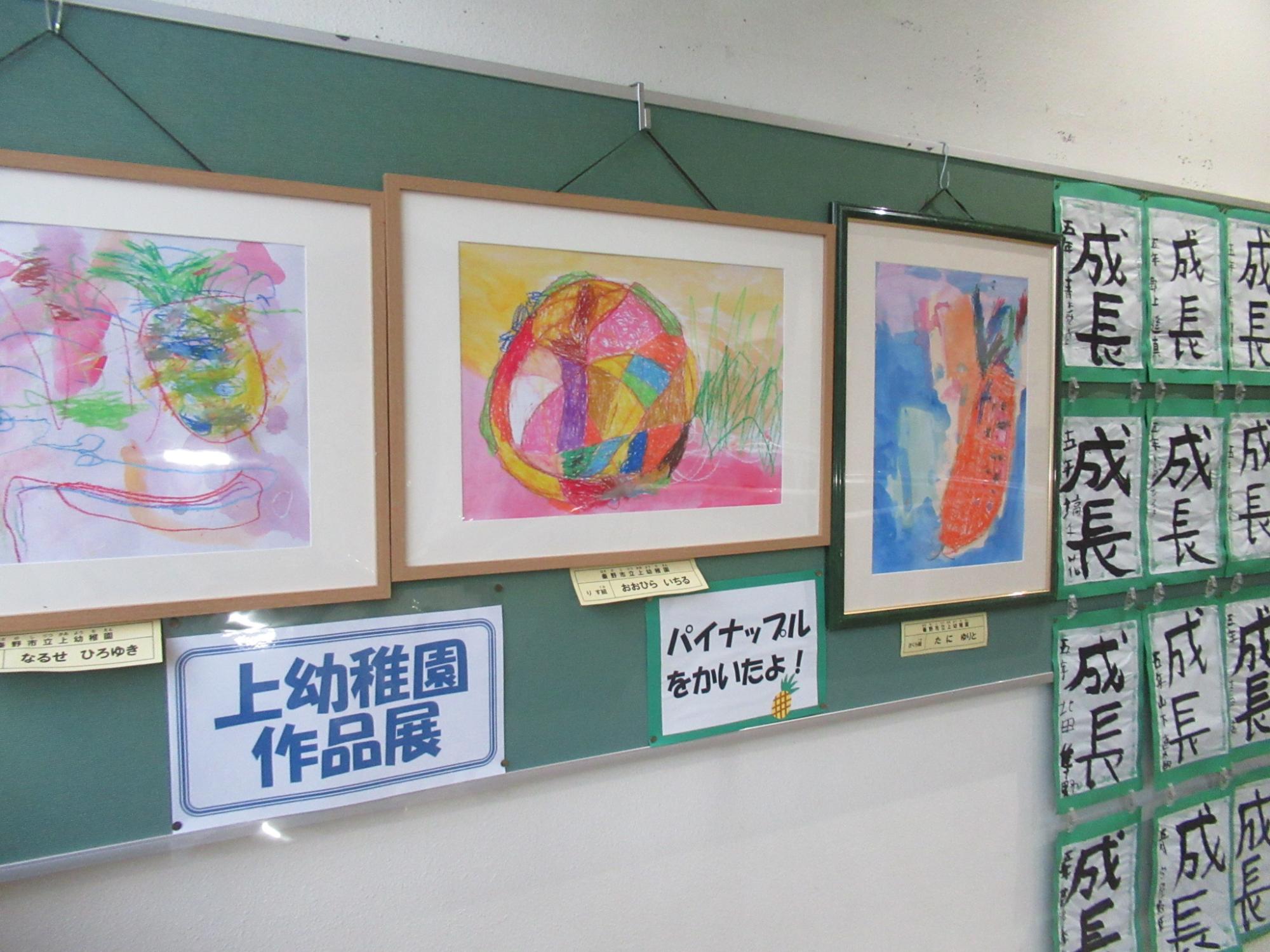 上幼稚園作品展と書かれた展示スペースに子どもたちが描いた額縁入りのカラフルな絵画が並び、右側の壁には「成長」と書かれた習字の作品が多数掲示されている写真