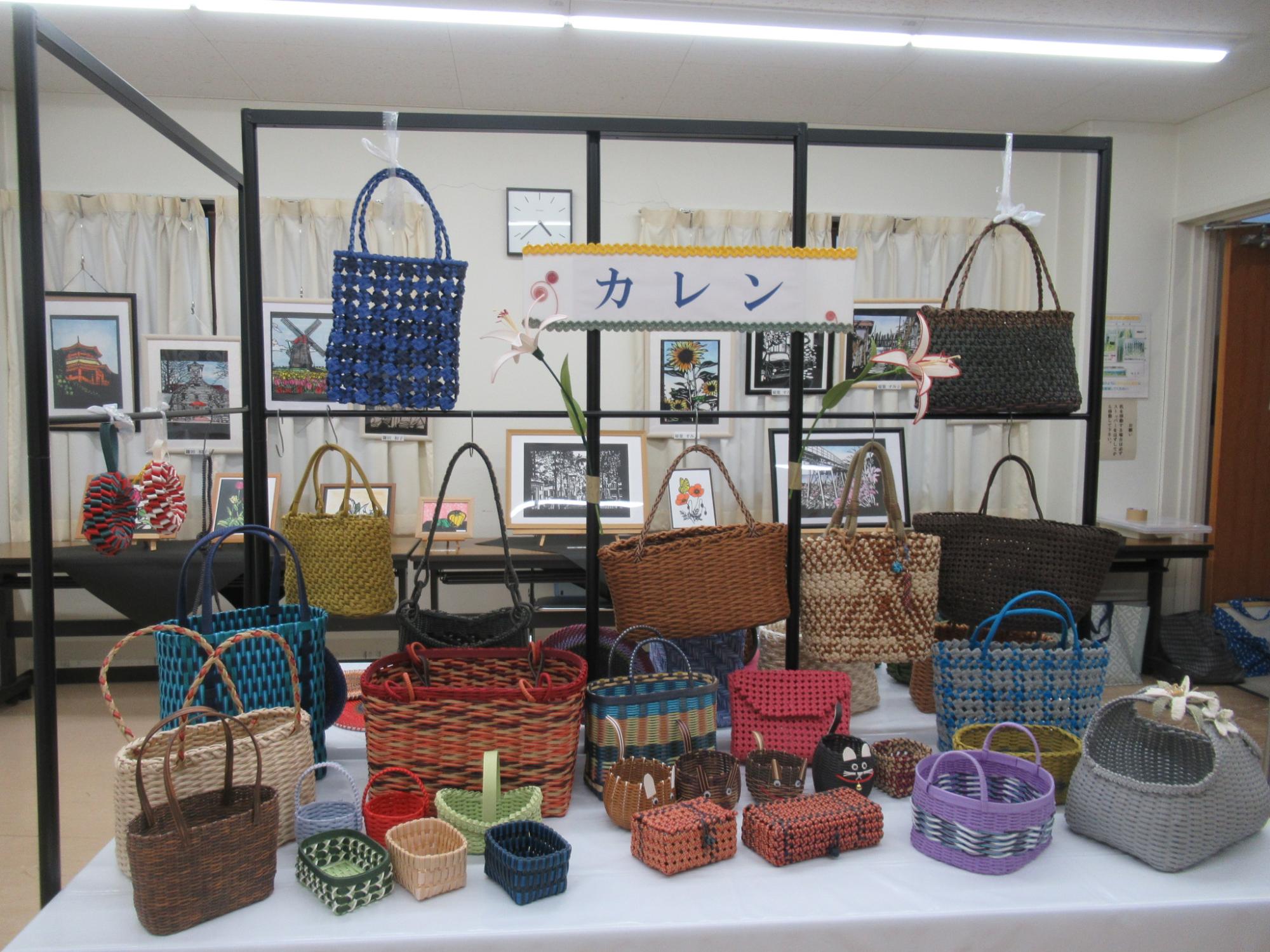 展示会場で、クラフトバンドなどで作られた多数のカラフルな編みカゴやバッグが展示され、背後の棚には「カレン」という名前の看板が掛けられている写真