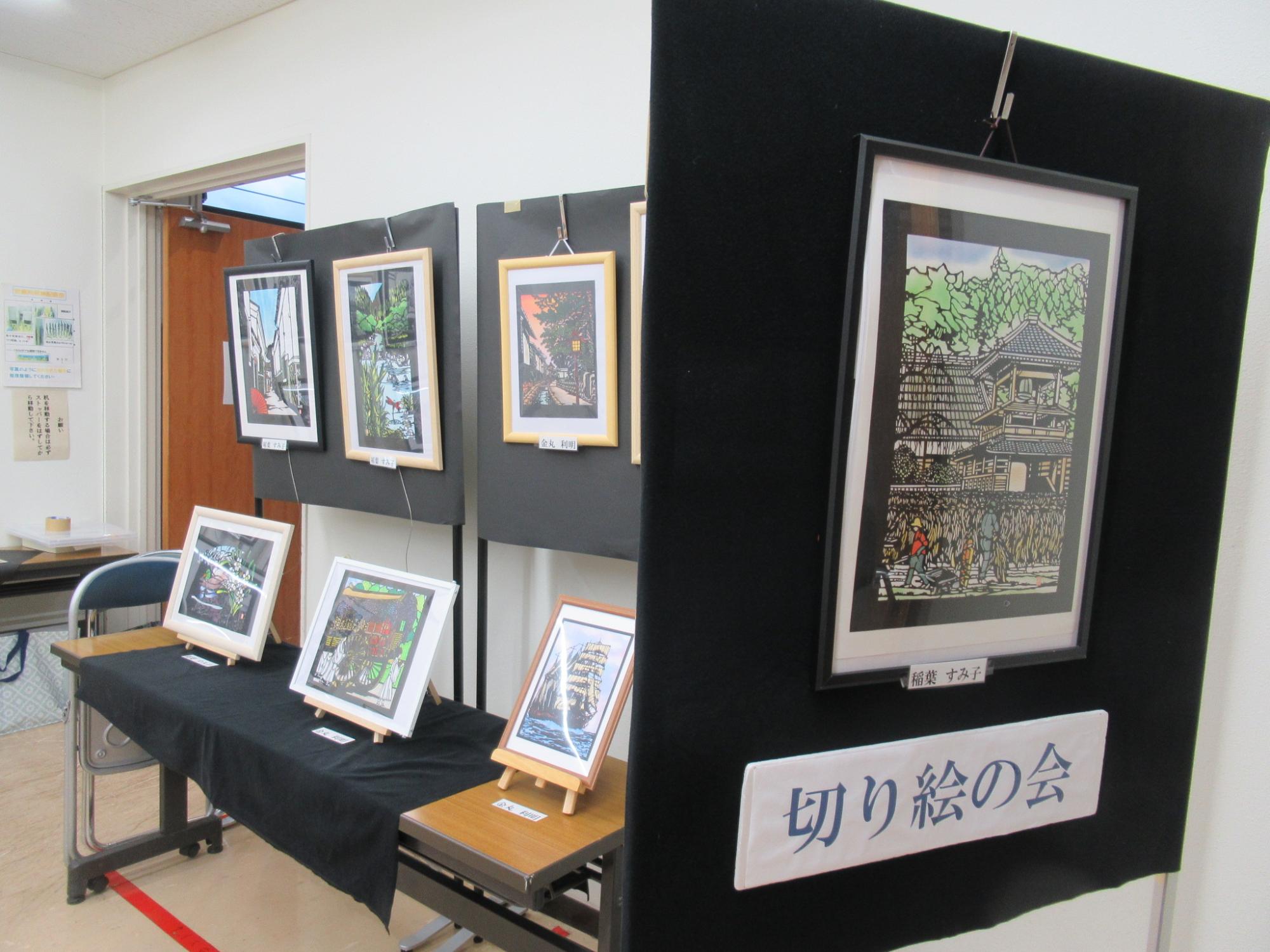 展示会場で、「切り絵の会」と書かれたボードやテーブルに、風景や建物などを細かく表現した複数の額装された切り絵作品が展示されている様子の写真