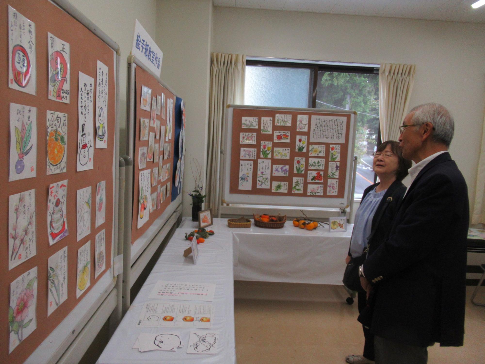 展示室で、壁一面にイラストが描かれた絵手紙作品が多数掲示され、テーブルの上には柿などが飾られており、男女2人がその作品を鑑賞している写真