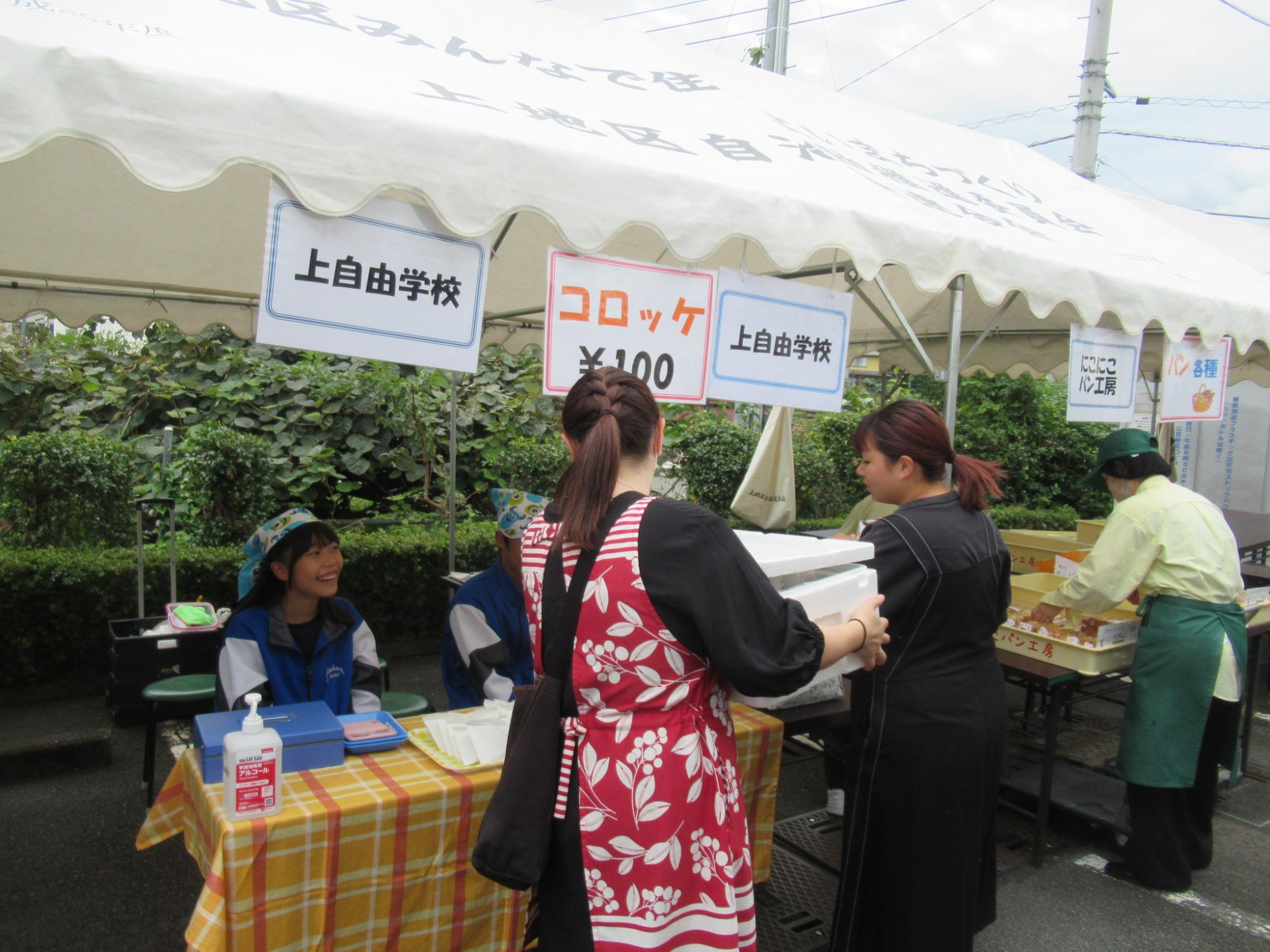 「コロッケ」の看板が下がった白いテントの下で椅子に座った女性が、店の前で足を止めた女性たちと会話をしている様子の写真