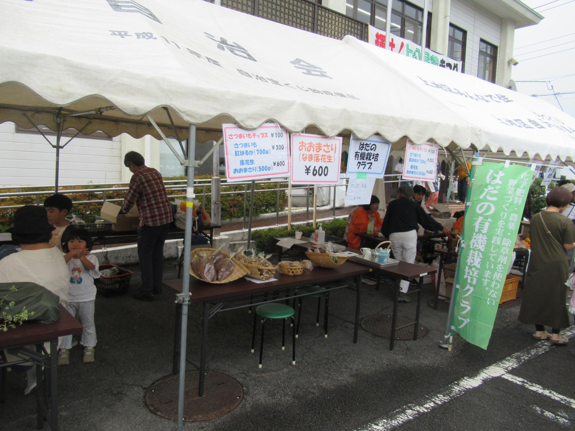 白いテントに商品名や価格が書かれたポップが多数掲示されており、中には商品が並んだテーブルがあり、複数の人がいる様子の写真