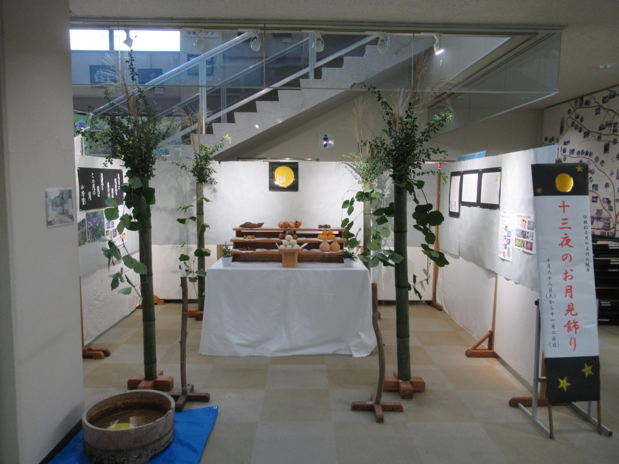 エントランスホールに、団子や季節の果物などの供え物が並べられ白い布をかけた台があり、両脇には植物の飾りと、「十三夜のお月見飾り」と書かれた幟が立てられている写真
