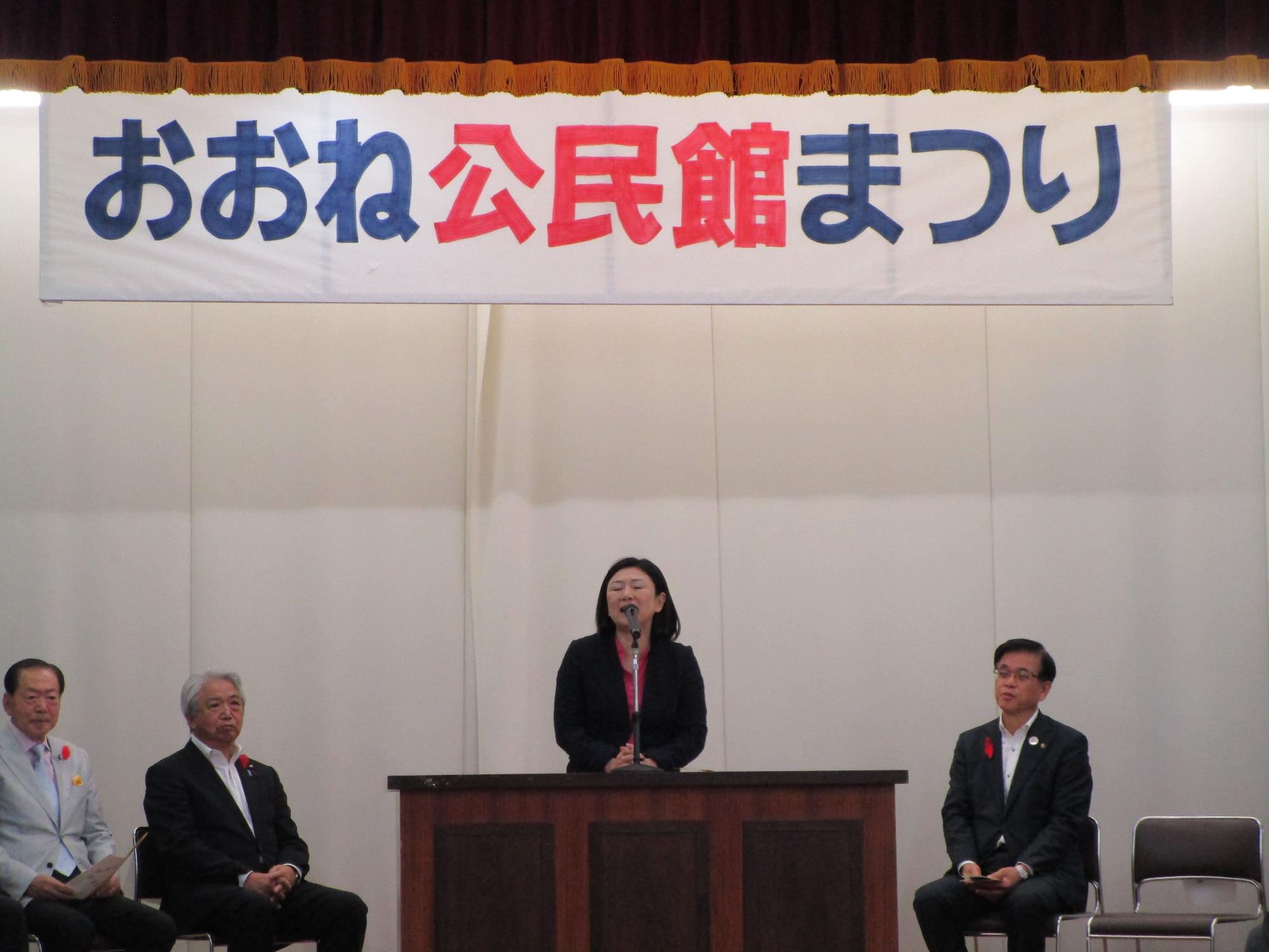 「おおね公民館まつり」と書かれた横断幕の下、ステージ上の演台でスーツ姿の女性がマイクて挨拶をしており、左右には数名の来賓らしき男性が着席している様子を捉えた写真