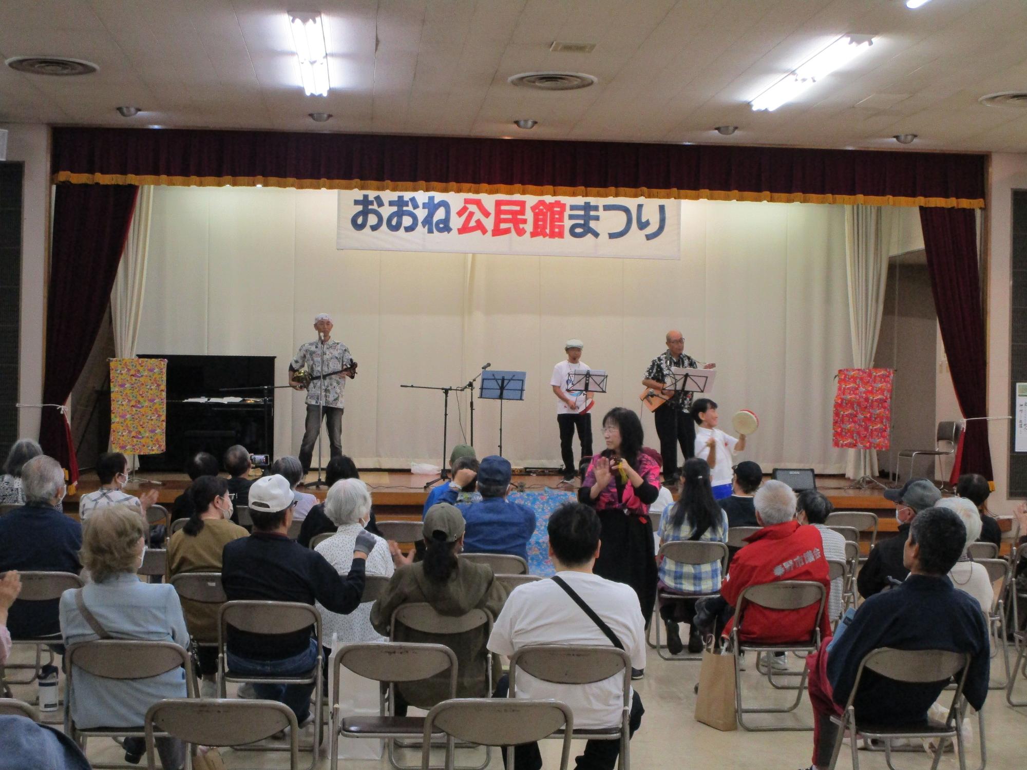 「おおね公民館まつり」と書かれた横断幕が掲げられたステージ上で3名がウクレレのような楽器を演奏し、ステージ下でタンバリンを叩いたり、客席の横を歩いて手拍子をしてしている様子の写真