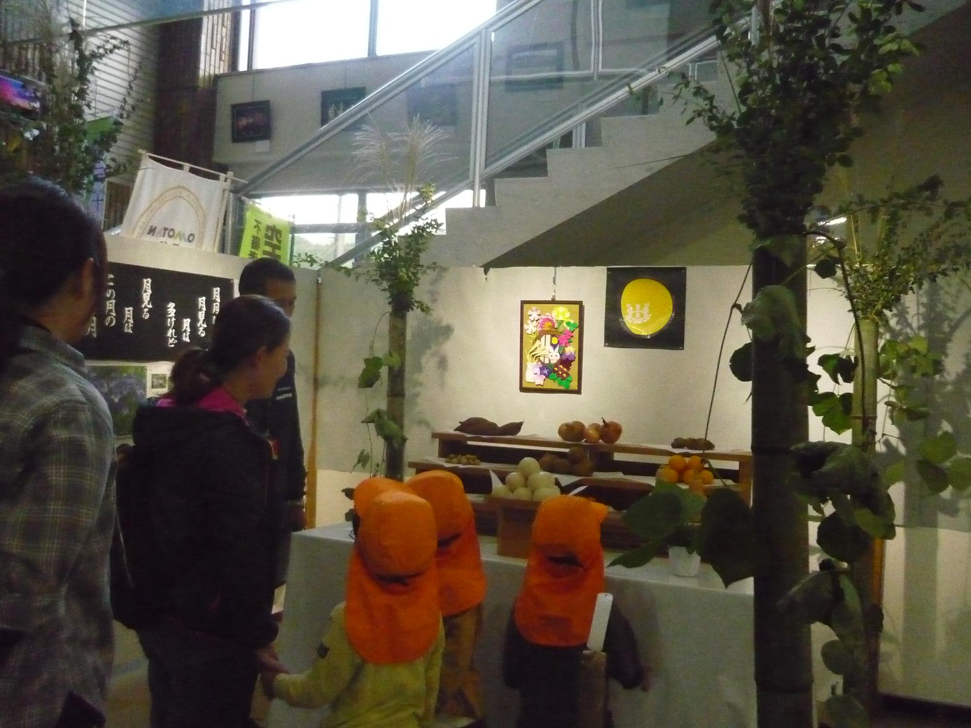 エントランスホールに設けられた十三夜のお月見飾りの展示スペースにオレンジ色の帽子を被った3人の小さな子どもたちと大人が集まっている写真