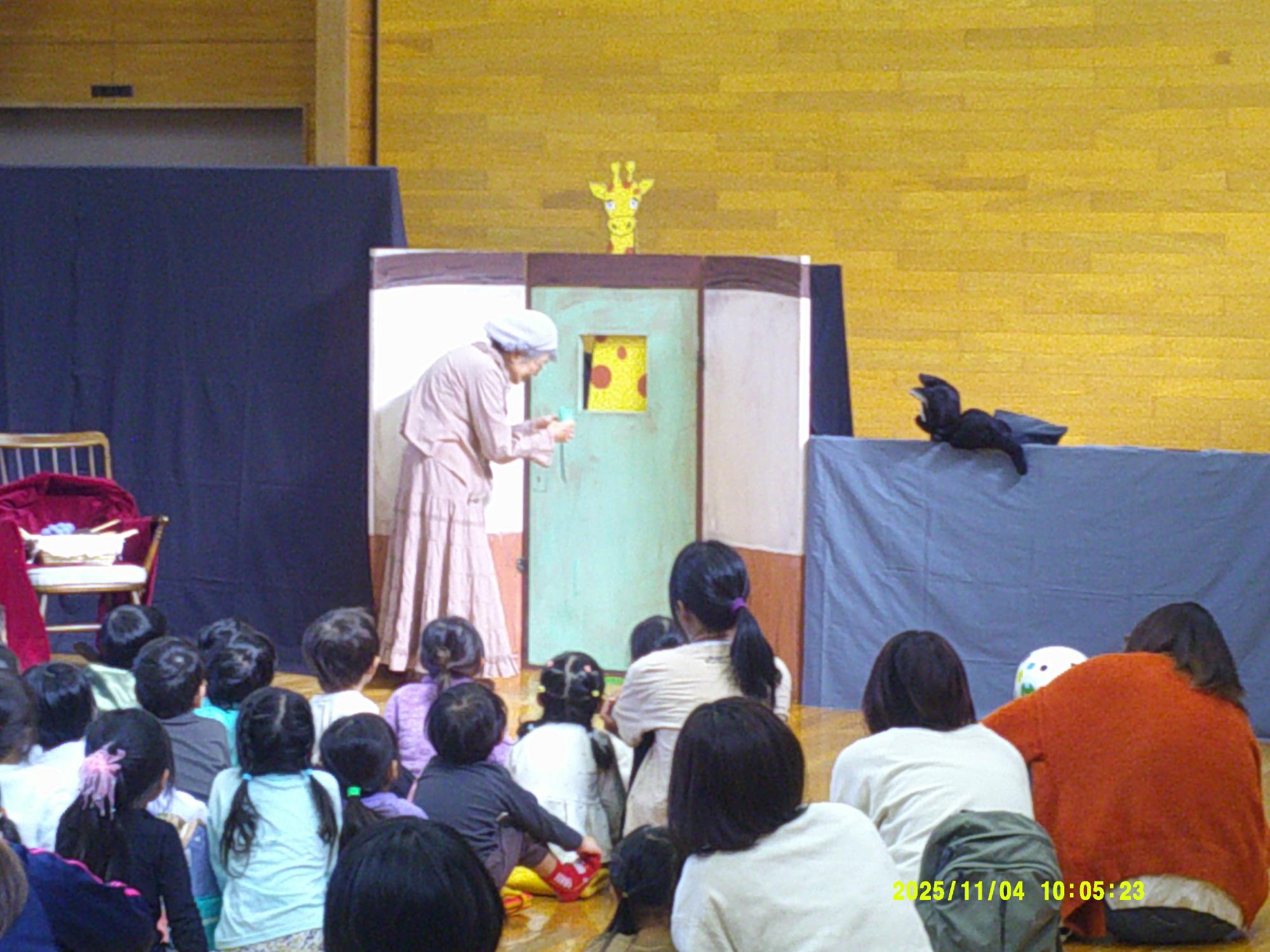 ホールのような広い空間で、エプロン姿の女性が人形劇のセットの前で演技しているのを床に座って真剣に見つめる多くの子どもたちや親子連れが鑑賞している写真