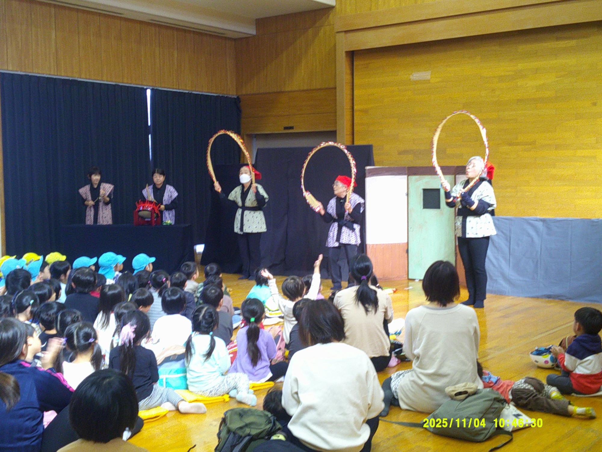 和服のような衣装を着て輪っかを持った5人の演者が黒い幕の前で舞台に立ち、床に座っている大勢の子供たちや大人たちがそれを見ている様子の写真