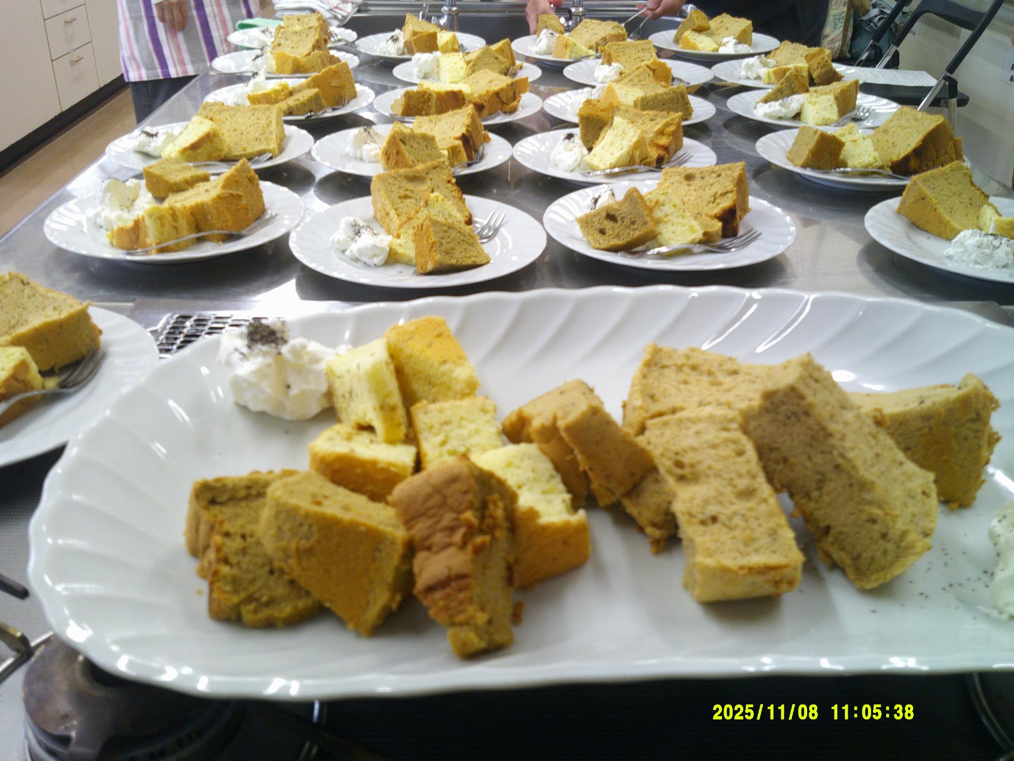 大きな長方形の白い皿に盛り付けられた茶色や薄い黄色のシフォンケーキまたはパウンドケーキの切り分けられた一切れとホイップクリームが、作業台の上に何皿も並べられている様子の写真