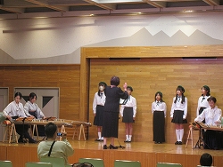 ステージ上にて、指導者らしき女性が指揮を執り、右側には制服姿の生徒たちが立ち、左側では琴を演奏している発表会の様子の写真