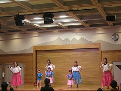 山脈が描かれたステージ背景の前で、鮮やかなピンクや青の衣装を着た数人の女性と小さな子供たちが、観客に向けてフラダンスを披露している様子の写真