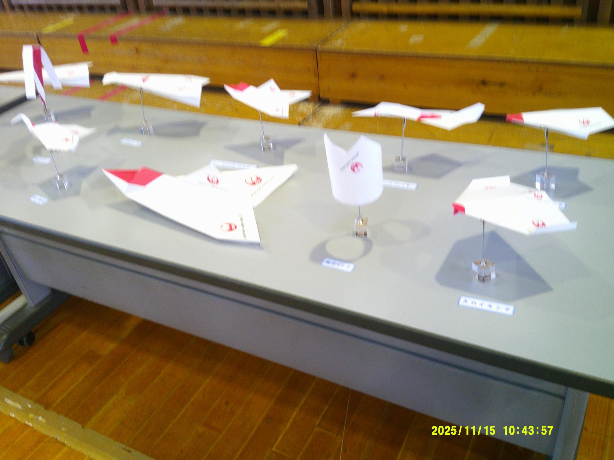 木目の床の体育館のような場所で、長机の上に白と赤を基調とした紙飛行機や紙の模型が多数展示されている写真