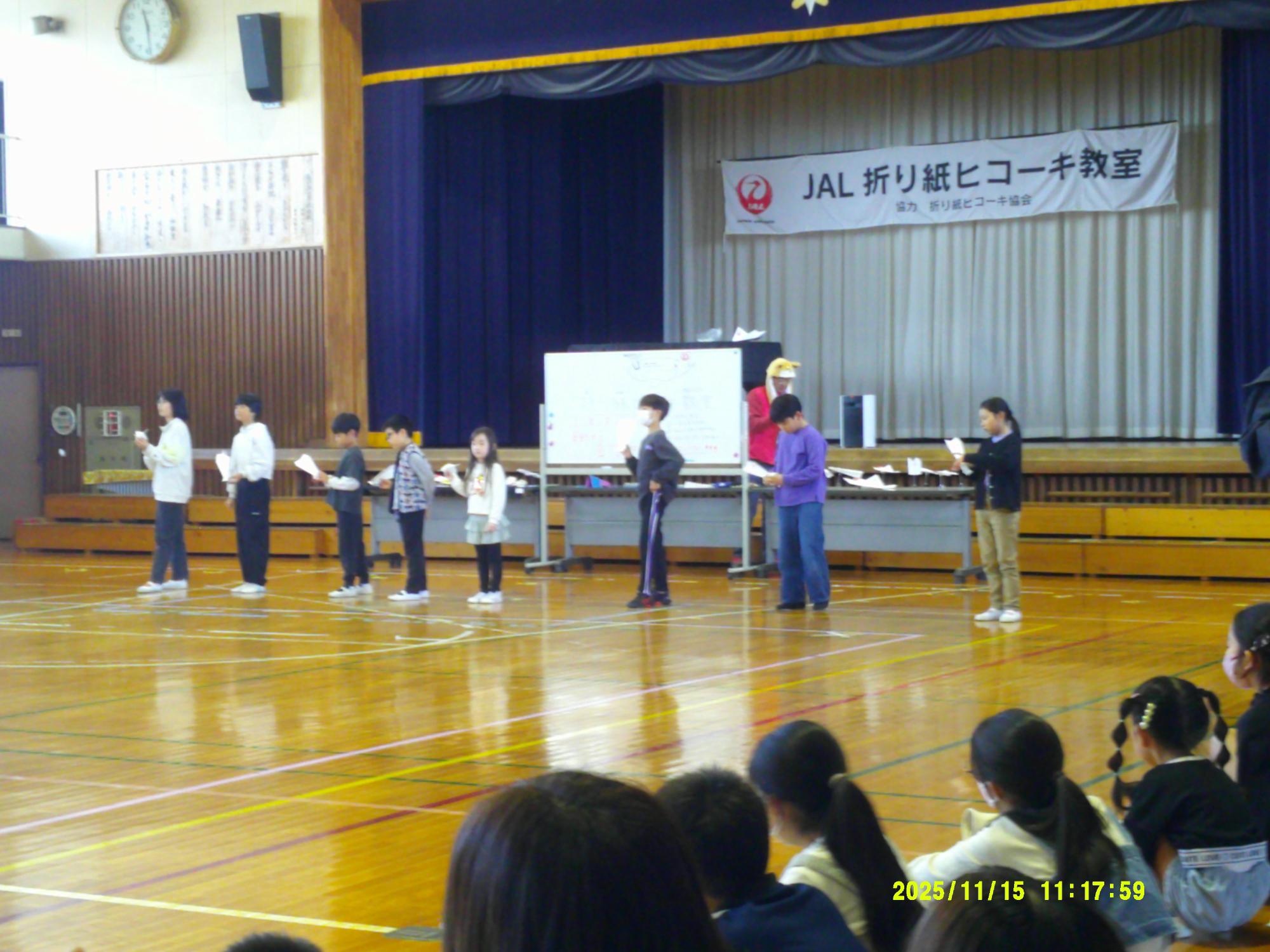 体育館のステージ前で、JAL折り紙ヒコーキ教室のバナーの下、数人の子供たちが紙飛行機を持ち、指導者らしき人物から説明を受けている様子を観客席側から写した写真
