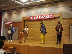 舞台の上に横一列に並んだ、和傘をさし太鼓を持つ男性、大太鼓を叩く着物を着た女性、サックスを演奏する男性、アコーディオンを弾く男性の4人の演者が演奏を行っている写真