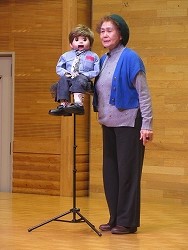 青いカーディガンを羽織った女性が、スタンドの上に固定された少年年型人形の横に立ち腹話術を披露している写真