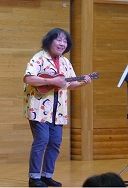 明るい表情の女性が、キャラクターの柄が入った黄色いシャツとジーンズを着用し、ウクレレを弾きながら舞台の上で演奏を披露している様子を捉えた写真