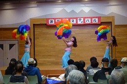 水色のスカートと肌色のトップスを着用した3人の女性が、鮮やかな虹色のファンベールを使い、ベリーダンスのような華やかな舞踊を舞台上で披露している写真