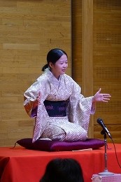 薄い色合いの着物を着用した女性が、紫色の座布団の上に正座し、小さなマイクに向かって両手を使いながら、落語を披露している様子の写真