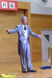 光沢のある薄いグレーの燕尾服を着用した男性が、両手に持った二つの輪を使い、観客に向かってマジックかリングを使ったパフォーマンスを披露している写真