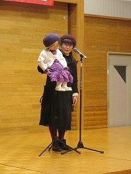 黒い服装にベレー帽をかぶった女性が、同じくベレー帽と紫のスカートを履いた人形を抱きかかえ、マイクの前に立って舞台上で腹話術を行っている写真