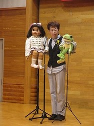 短髪で眼鏡をかけた演者が、右手に頭にピンク色のリボンをつけた女の子の人形、左手にカエルのパペットを持ち腹話術を披露している写真