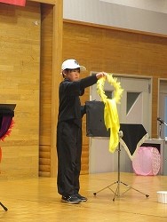 舞台上のマイクスタンド付近で、白いキャップをかぶった黒いジャージ姿の子どもが、黄色い長い布と輪を使い、手品を披露している瞬間を捉えた写真