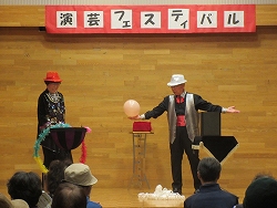 白いハットとシルバーのベスト、蝶ネクタイの男性マジシャンが、横にいる赤いハットの女性とともに、ピンクの風船を使った手品を舞台の上で観客に披露している写真