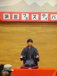 紺色の着物を着用した男性が、舞台の中央に置かれた赤い座布団の上に正座し、低めのマイクに向かって両手を広げながら落語を披露している写真