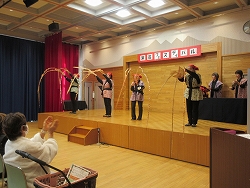 赤い頭巾を被り、和風の衣装を身につけた4人の演者が、長い竹のような棒を大きく振り上げる南京玉すだれを舞台上で披露しており、観客が拍手している写真