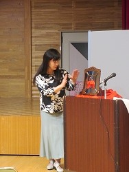 黒と白の柄のトップスとミントグリーンのロングスカートを着用した長い黒髪の女性が、演台に置かれた砂時計に両手をかざして、手品を披露している写真