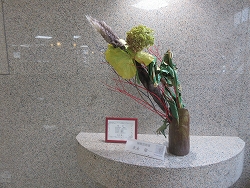 茶色の木製の花器に生けられた、緑色の大きな葉と赤い枝、黄色や紫の素材を組み合わせた現代的な生け花作品の写真