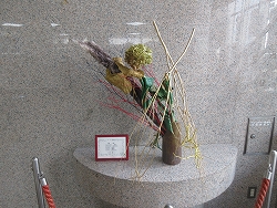 茶色の筒状の花器に、金色や赤色の細い枝、濃い緑の布状の素材、そして紫や黄色の実のような素材を立体的に組み合わせた生け花の作品が展示されている写真