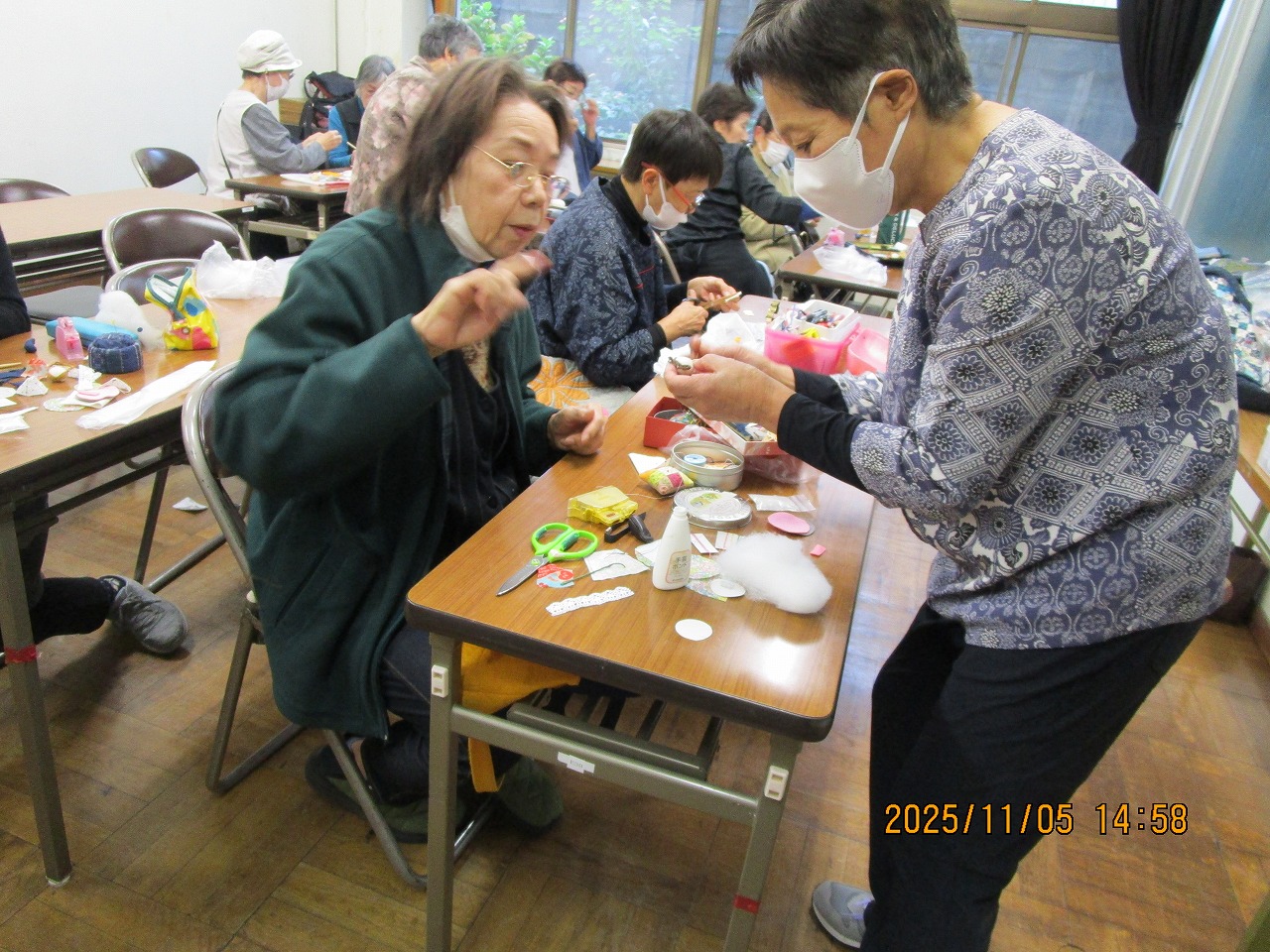 長机にハサミや綿などの材料を広げて手芸に取り組む眼鏡をかけた女性に対し、マスク姿の女性が立って手元を見せながら指導している写真