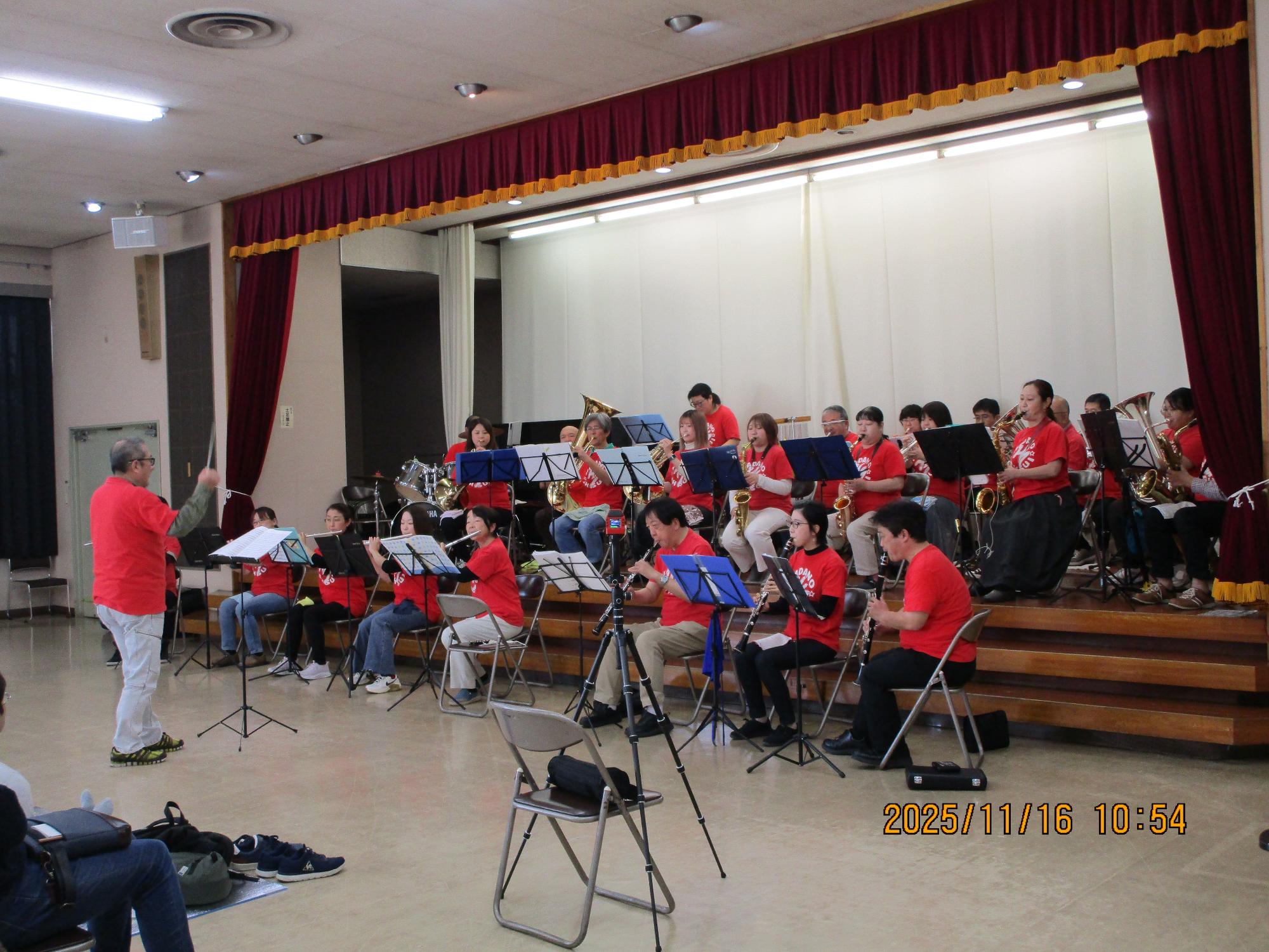 ステージ上で赤いTシャツを着た吹奏楽団が指揮者に合わせてフルートやサックスなどの管楽器を演奏している写真