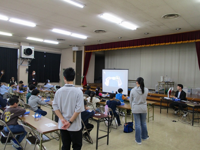 広い会場に並べられた長机で大勢の子供たちが工作に取り組み、前方のスクリーンに映し出された手元の映像を参考にしながら作業を進め、その様子を数人の大人たちが見守っているかながわリレー教室の写真