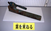 「葉を束ねる」と書かれた札が置かれ、長細い金属の棒と、先端のレバーのような部分が錆びて変色した、古い道具が台の上に置かれている写真