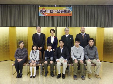 「親子川柳大会表彰式」の横断幕の下に設置した金屏風の前で、前列の椅子に3組の親子が座り、後列にスーツ姿の男女4人が並んで立って写した記念写真