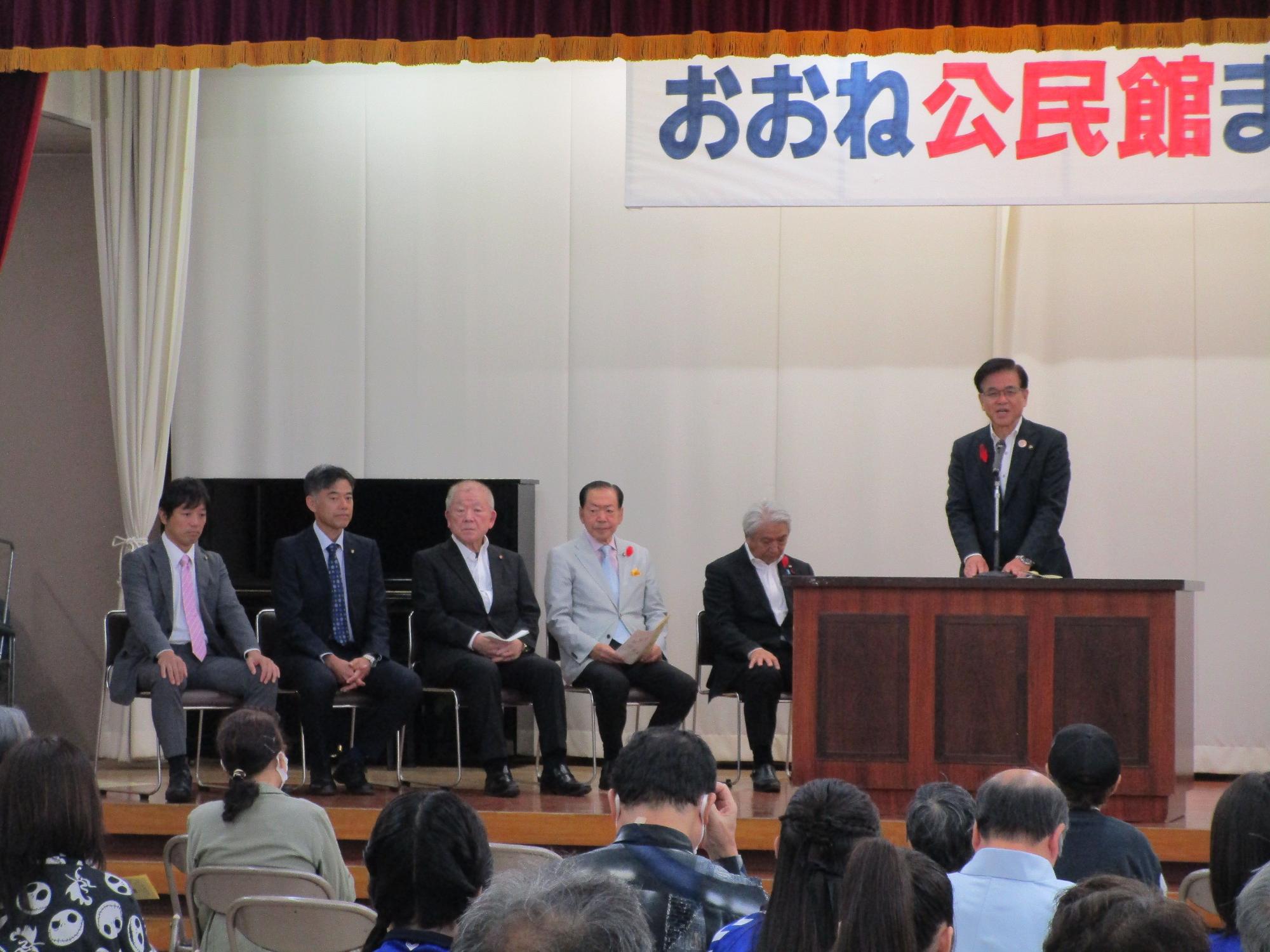 公民館のような会場のステージに「おおね公民館まつり」と書かれた横断幕が掲げられ、壇上の演台で高橋昌和秦野市長が挨拶をしている様子を、聴衆の後方から捉えた写真